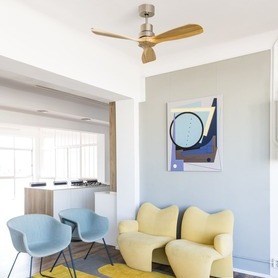 Salon lumineux avec fauteuils bleus et jaunes et un ventilateur au plafond.