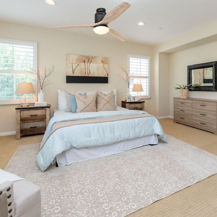 Ventilateur de plafond au-dessus d’un lit dans une chambre lumineuse et meublée avec table de chevet et commode.